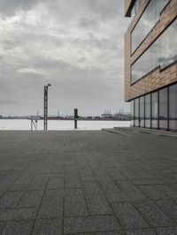 an empty sidewalk next to the water in front of a building with large glass windows