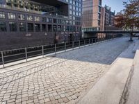 a bench on a brick sidewalk near a bridge with a train going over it and some buildings and trees on one side