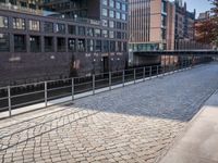 a bench on a brick sidewalk near a bridge with a train going over it and some buildings and trees on one side