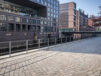 a bench on a brick sidewalk near a bridge with a train going over it and some buildings and trees on one side