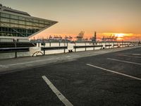 some cars parked near water and a building with some boats in the background at dusk