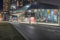 the view of a city from across the street at night showing buildings and a bus stop