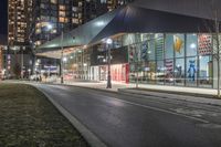 the view of a city from across the street at night showing buildings and a bus stop