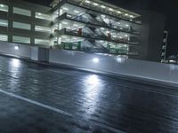 a dark street next to a building at night with a lit up parking garage behind it