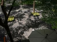 an umbrella sits in the shade as pedestrians walk by in a park area with trees