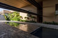 a pool in an open building with lots of plants in the background, and a water feature, along the concrete wall that connects to the swimming pool and large stone steps leading the outdoor patio