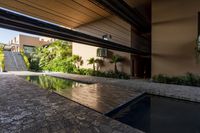 a pool in an open building with lots of plants in the background, and a water feature, along the concrete wall that connects to the swimming pool and large stone steps leading the outdoor patio