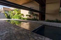 a pool in an open building with lots of plants in the background, and a water feature, along the concrete wall that connects to the swimming pool and large stone steps leading the outdoor patio