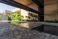 a pool in an open building with lots of plants in the background, and a water feature, along the concrete wall that connects to the swimming pool and large stone steps leading the outdoor patio