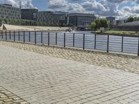 a paved sidewalk near a river and some water and buildings in the distance behind it