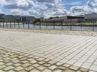 a paved sidewalk near a river and some water and buildings in the distance behind it