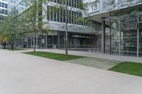 the sidewalk in front of a building has grass, benches, and trees on it