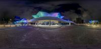 a large modern architecture structure lit up in colors of purple and blue in a circular space with stone walkway