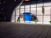 some people walking in a building near a large glass window at night by some pots