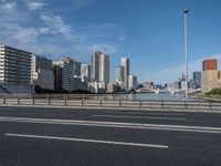 a road with tall buildings in the background and water under a blue sky near it