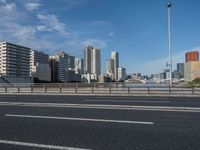 a road with tall buildings in the background and water under a blue sky near it