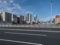 a road with tall buildings in the background and water under a blue sky near it