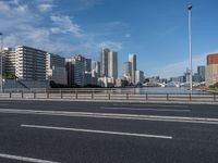 a road with tall buildings in the background and water under a blue sky near it