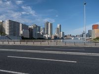 a road with tall buildings in the background and water under a blue sky near it