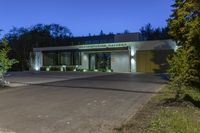 a green and white building next to a parking lot at night with trees lining the sidewalk