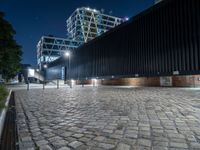 a brick walkway with lights shining down on the ground and the building at night in the distance