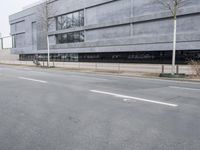 this is an empty road near a gray building with a building in the background and trees