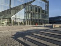 a walkway near a large glass building with stairs and benches outside and people walking past