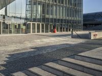 a walkway near a large glass building with stairs and benches outside and people walking past