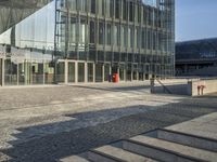 a walkway near a large glass building with stairs and benches outside and people walking past