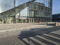 a walkway near a large glass building with stairs and benches outside and people walking past