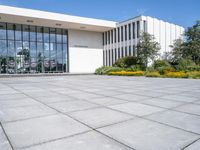 a white building with a glass entrance next to a row of yellow flowers on each side of the entrance is surrounded by green grass and trees