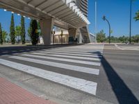 a car sitting at an empty street underneath a overpass overhang that contains a train stop, and cars and a pedestrian