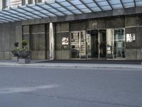 a street is lined with cement and glass doors of a building that has columns, plants and a few round bushes on one side and windows