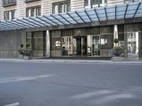 a street is lined with cement and glass doors of a building that has columns, plants and a few round bushes on one side and windows