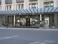 a street is lined with cement and glass doors of a building that has columns, plants and a few round bushes on one side and windows