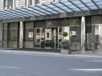 a street is lined with cement and glass doors of a building that has columns, plants and a few round bushes on one side and windows