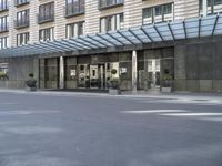 a street is lined with cement and glass doors of a building that has columns, plants and a few round bushes on one side and windows