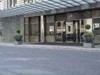 a street is lined with cement and glass doors of a building that has columns, plants and a few round bushes on one side and windows