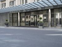 a street is lined with cement and glass doors of a building that has columns, plants and a few round bushes on one side and windows