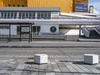 a walkway with a bench outside a modern building and yellow facade in the background, a clock on a post