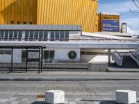 a walkway with a bench outside a modern building and yellow facade in the background, a clock on a post