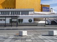 a walkway with a bench outside a modern building and yellow facade in the background, a clock on a post