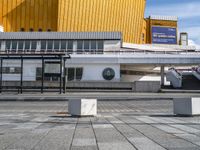 a walkway with a bench outside a modern building and yellow facade in the background, a clock on a post