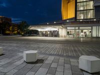 an empty plaza and a lit up building at night with two square stone benches in front