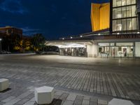 an empty plaza and a lit up building at night with two square stone benches in front