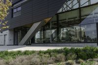 black clad, modern building with glass front and green bushes in front of it and the sidewalk next to it