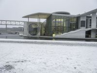 a large glass and stone building next to a river with snow on the ground and a person walking across the bridge