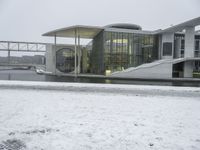 a large glass and stone building next to a river with snow on the ground and a person walking across the bridge