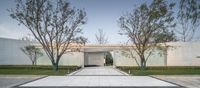 the entrance to a large modern building with several trees and bushes outside of it as well as a pathway between two large, rectangular white, glass walled walls