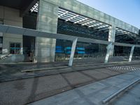 a bench sitting in front of a glass building on top of stone pavements below a metal ceiling